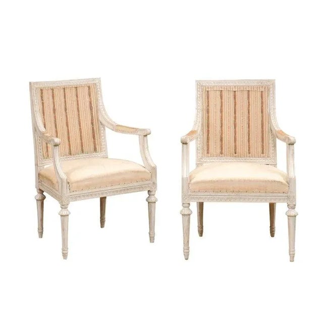 A pair of Swedish Gustavian style painted wood armchairs from the early 20th century, with scrolling arms, carved...