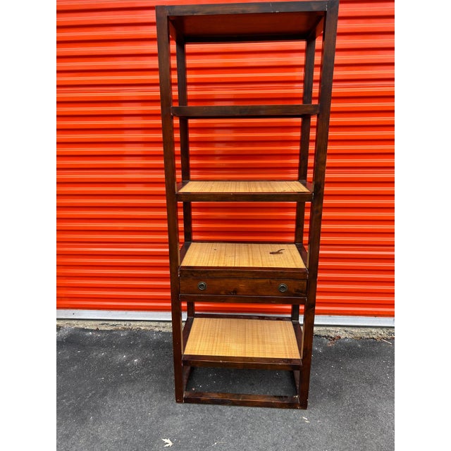 This tall, multi-level shelving unit features a clean, architectural frame crafted in dark-stained wood with woven cane...