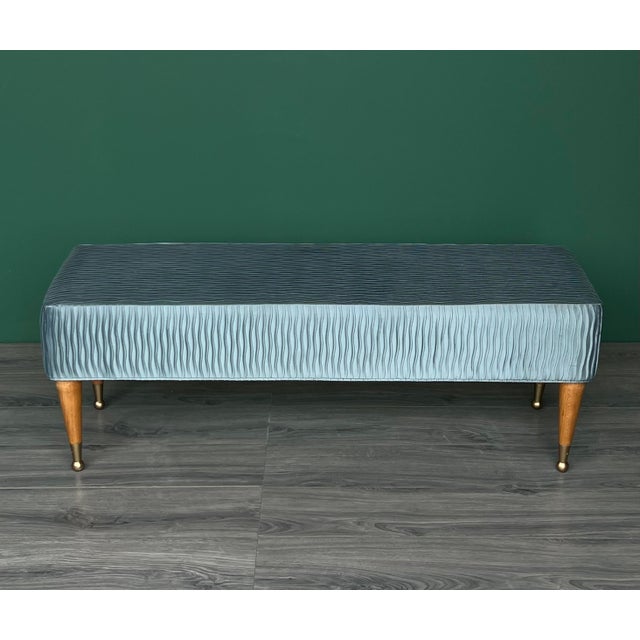 This Mid-Century bench is newly upholstered in a turquoise pleaded satin textile and supported by walnut tapered legs....