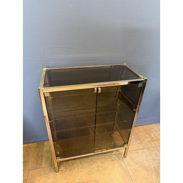 This brass and smoked glass display cabinet rests on four square feet. It has four shelves: three interior shelves and a...