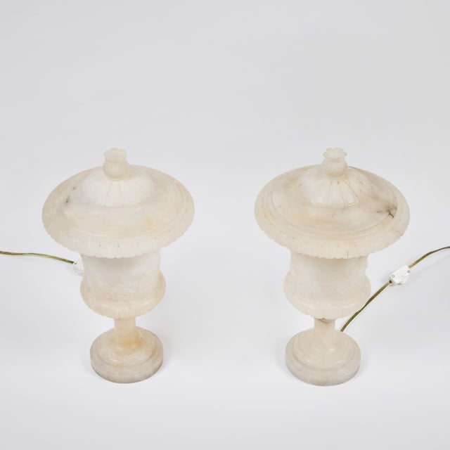 Pair of hand-carved alabaster urns as lamps, featuring putti and flower reliefs in body and scalloped edged lids with...