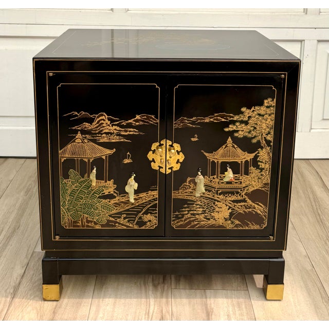 This vintage, mid-20th century black lacquer Chinoiserie nightstand cabinet features richly detailed gilt scenic...