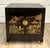 This vintage, mid-20th century black lacquer Chinoiserie nightstand cabinet features richly detailed gilt scenic...