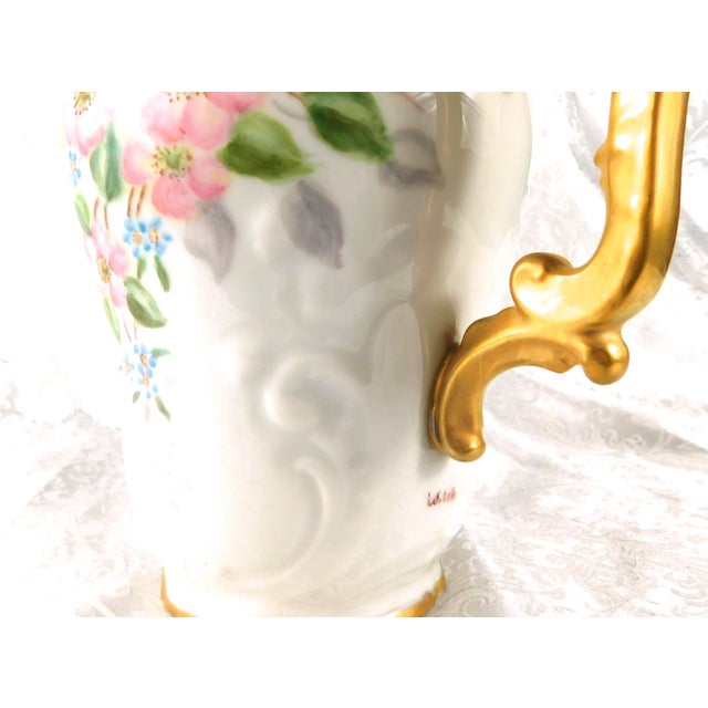 This is a white unmarked, hand painted and signed chocolate pot or teapot. It has pink flowers, smaller blue flowers and...