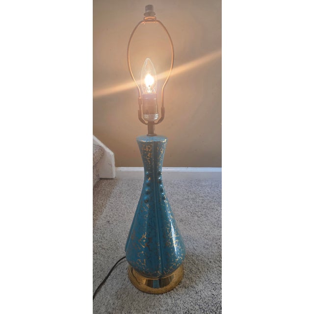 A stunning turquoise and gold Mid-Century porcelain lamp. This lamp is 25" tall, 6" wide. The surface features a varied,...