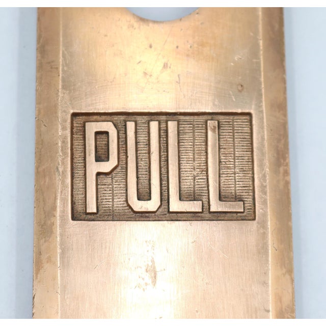 This vintage commercial brass door pull features a rectangular back plate with a center mounted arched pull handle and a...