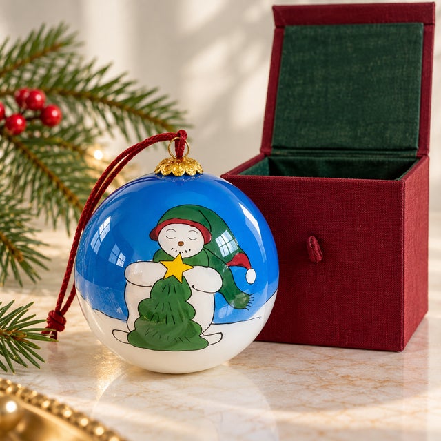 Delightful vintage glass Christmas ornament featuring a whimsical snowman decorating a Christmas tree beneath a vivid blue...