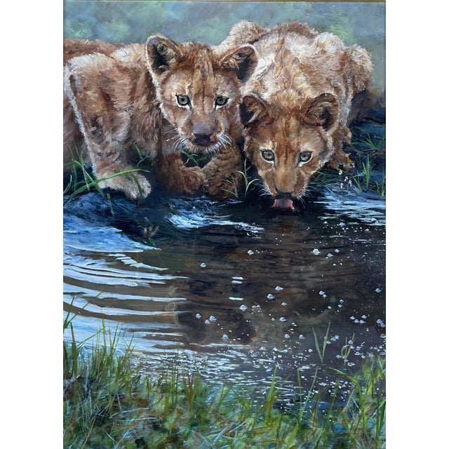 This oil on canvas artwork depicts two young lions at a tranquil riverbank, their reflections shimmering in the water. The...