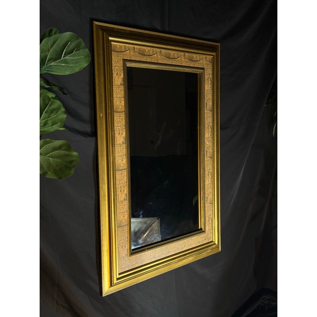 Ornate giltwood-style wall mirror with an Egyptian Revival inspired design. The rectangular frame features a wide gold-...