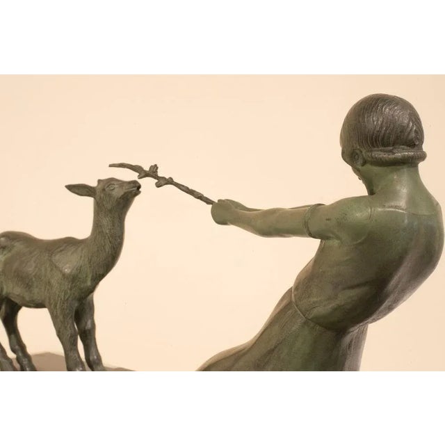 Metal sculpture on black and green rectangular marble base. A woman points a wand at a goat. The metal has a green patina...