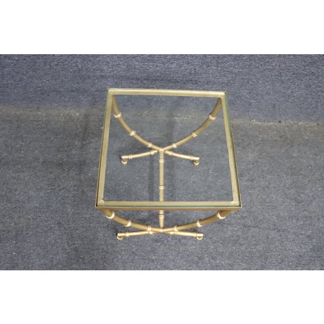 Square brass side table with a clear glass top and bamboo-style curved legs joined by an X-base. The warm gold finish...