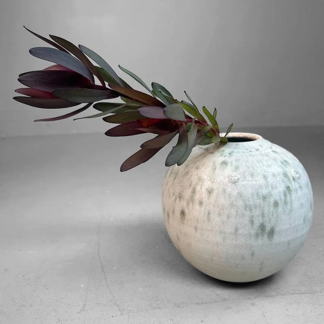 A restrained, spherical ikebana vase with a soft, almost cloud-like presence. The fine craquelure glaze shifts subtly from...