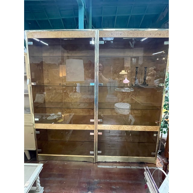 Two available- This Hollywood Regency display cabinet features smoked glass doors and shelves framed in polished brass...