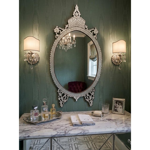 All white mother-of-pearl inlaid wall mirror featuring an elegant oval silhouette and ornate crown crest. The frame is...