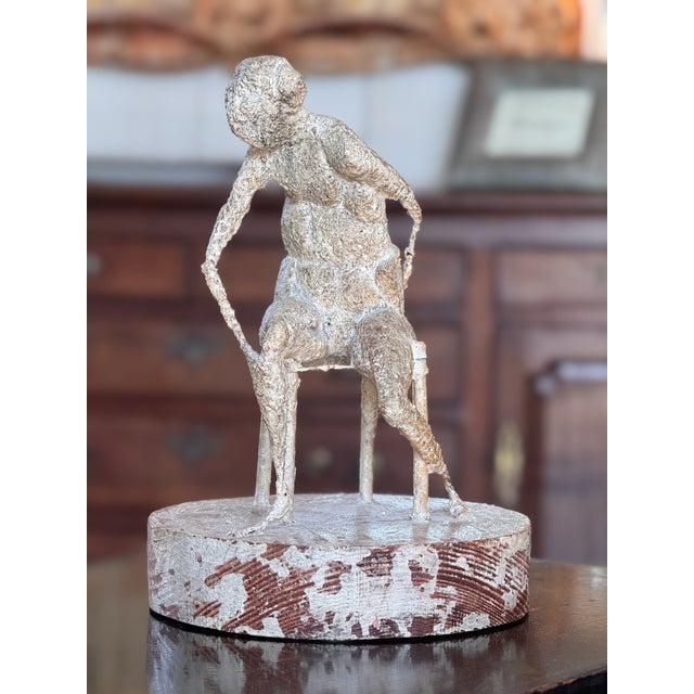 An abstract sculpture of a seated figure with elongated limbs, leaning to one side, the surface rough and plaster-like in...