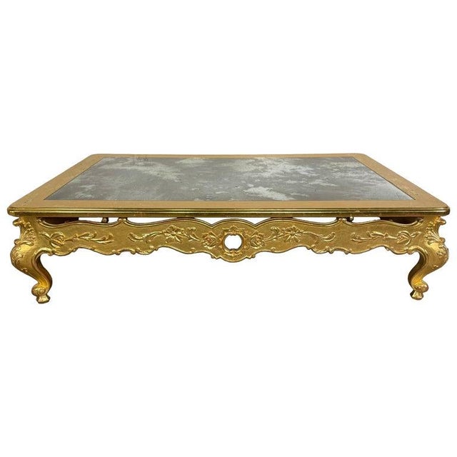 A striking Rococo-style giltwood coffee table featuring an inset antiqued mirrored top framed by a molded gilt edge. The...