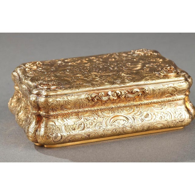 A cartouche-shaped gold box. The hinged lid, the base and the sides are chiseled with flowers and scrolls as well as...