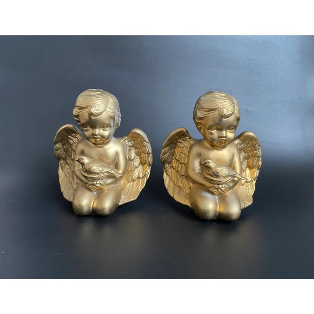 A pair of vintage bisque porcelain sculptures featuring gold-painted cherubs holding doves. These charming pieces are in...