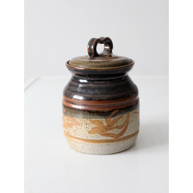 This vintage studio pottery jar features a rich, earthy palette with a dark brown top, a warm bronze band, and a speckled...