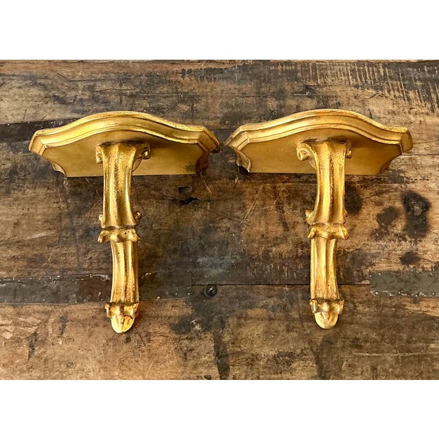 This exquisite pair of vintage gilt wall brackets brings a touch of opulent Neoclassical grace and Hollywood Regency...