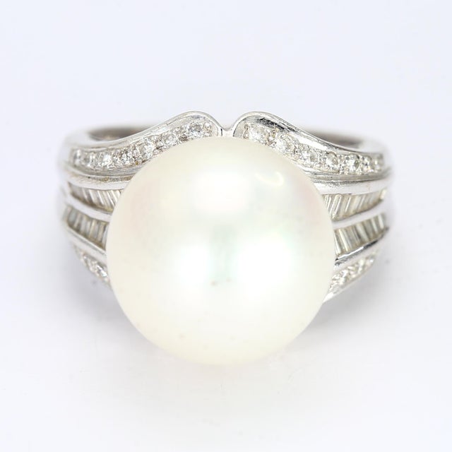 This is a large 18k white gold cocktail ring with a 14mm round pearl sat atop the ring while the accent diamonds seemingly...