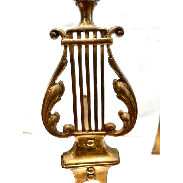 A refined pair of mid-20th-century brass andirons featuring elegant neoclassical lyre motifs and sculptural pineapple-...