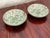 This is a pair of small porcelain plates. Each plate is set in a brass back. The fronts are painted with green floral...