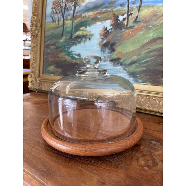 A mid-century modern cheese dome featuring a clear blown-glass cloche paired with a warm, solid teak wood base. While hard...