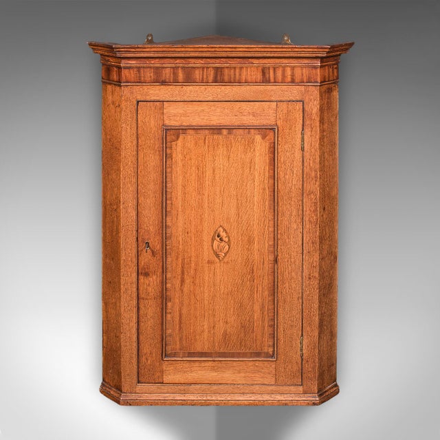 This is an antique mounted corner cabinet. An English, golden oak and mahogany wall cupboard with inlaid shell motif,...