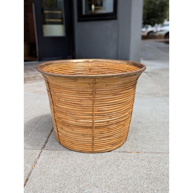 A cylindrical rattan vessel dating to the 1960s–1970s, reinforced with a rolled brass rim. The woven body provides visual...