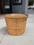 A cylindrical rattan vessel dating to the 1960s–1970s, reinforced with a rolled brass rim. The woven body provides visual...