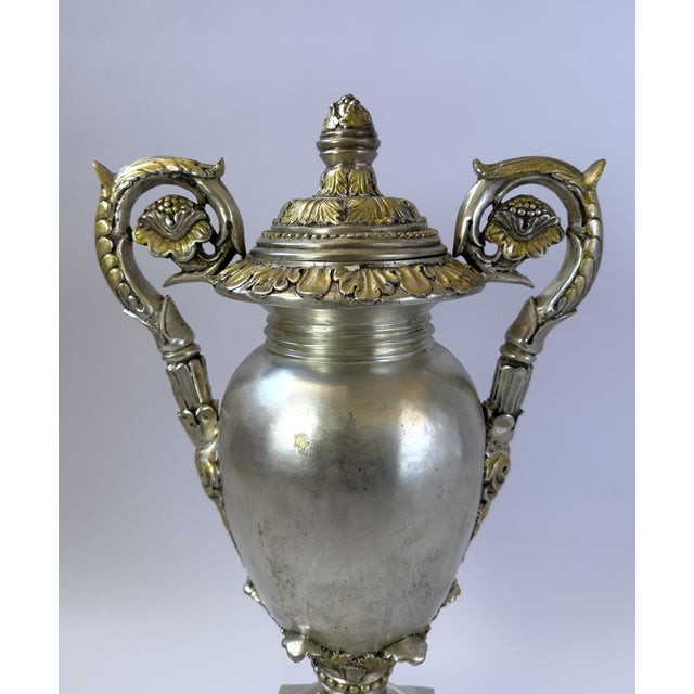 This impressive bronze urn features a brilliant silver finish with subtle golden highlights that accent its ornate...