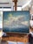 The artwork depicts a serene seascape, where soft, billowing clouds float above a tranquil ocean, illuminated by a gentle...