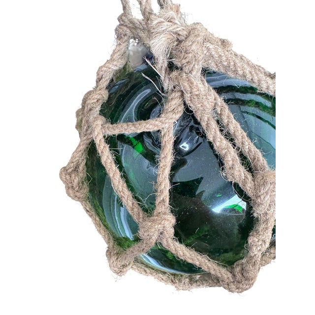 A hand-blown glass fishing float in a rich sea-green hue, hand-knotted into its original diamond-pattern jute rope netting...