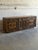 This vintage wooden sideboard features an expansive rectangular profile highlighted by a dramatic three-dimensional...