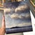This artwork presents a serene landscape, featuring a tranquil body of water with a distant island silhouetted against a...