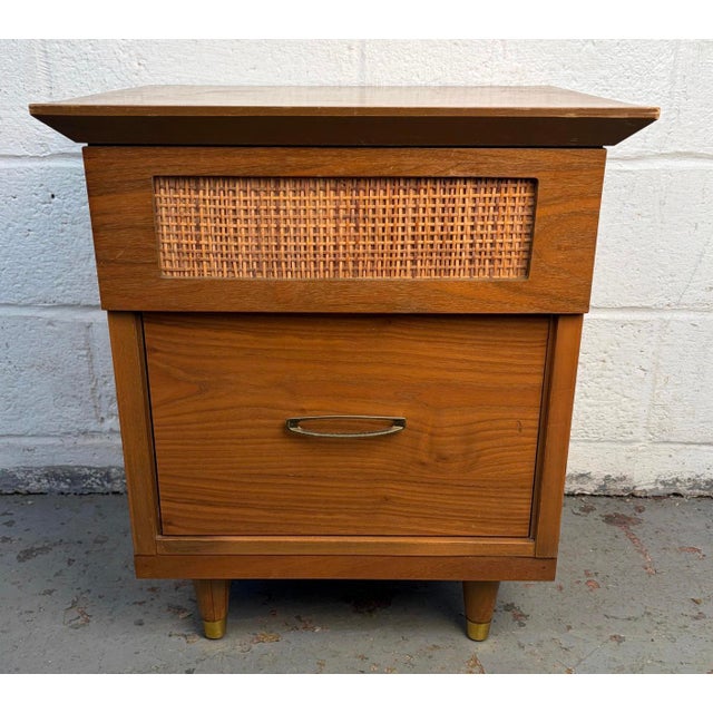 A charming mid-century modern walnut nightstand featuring a distinctive cane-faced drawer that adds texture and visual...