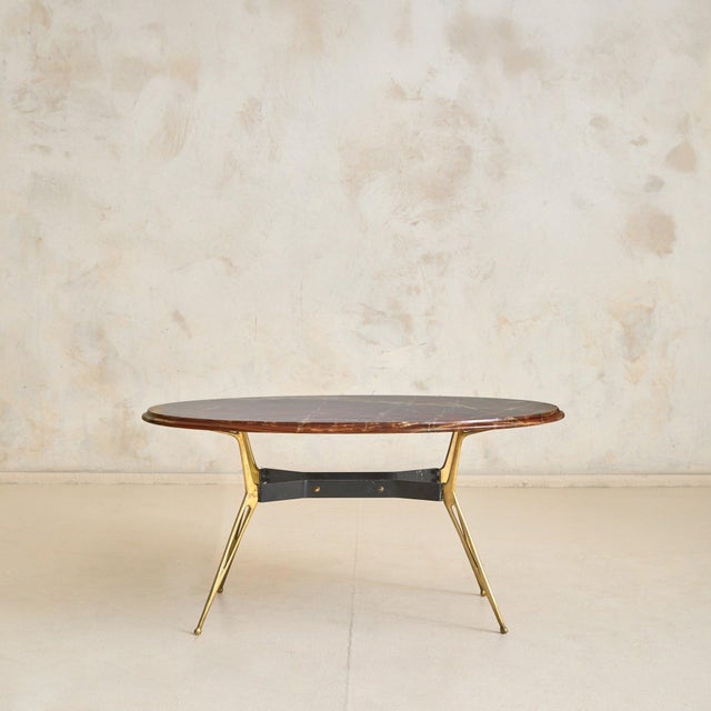 A 1950s Italian oval coffee table, defined by its richly veined onyx top and sculptural brass base. The oval top is carved...