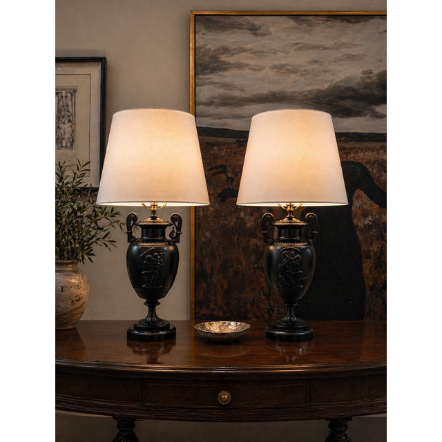 A refined pair of Neoclassical urn-form table lamps featuring sculptural figural reliefs and elegant scroll handles with...