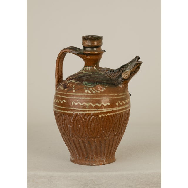 An earthenware ewer with an animal head spout with an open mouth and tooled fur texture. Consistent with rural pottery...