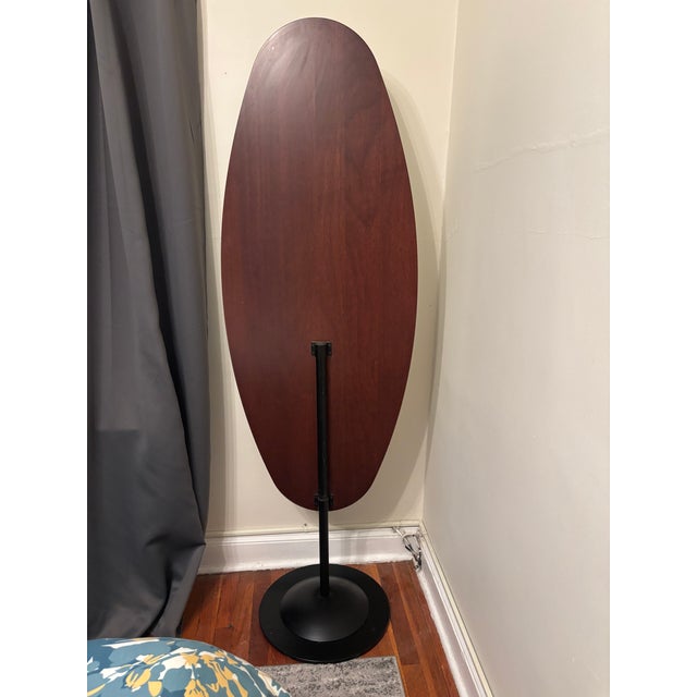 Striking sculptural floor mirror featuring an elongated oval silhouette, warm dark wood surround, and black metal pedestal...