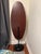 Striking sculptural floor mirror featuring an elongated oval silhouette, warm dark wood surround, and black metal pedestal...