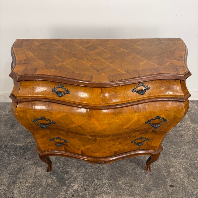 An Italian bombe-style chest of drawers. This piece features a parquet design with burlwood and mahogany and scrolled...