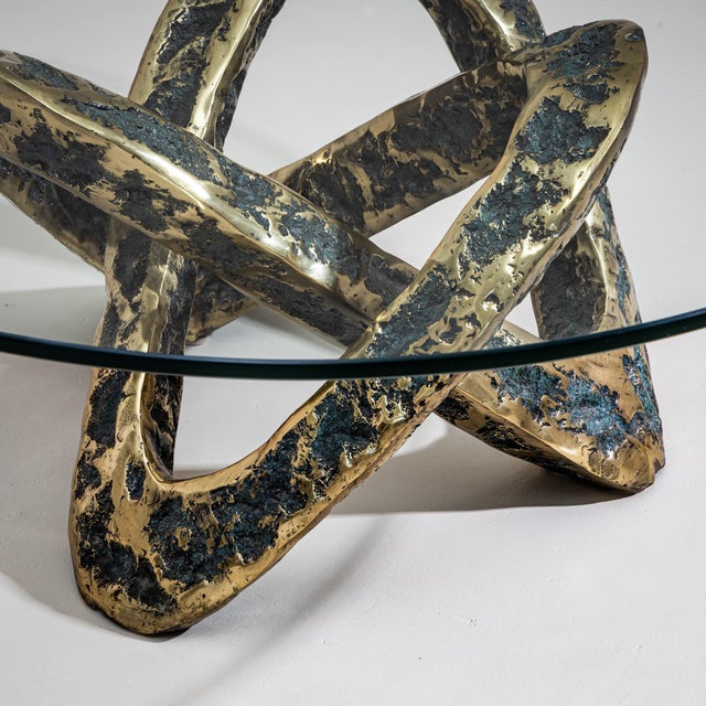 Round coffee table featuring a clear glass top and an intertwined bronze base. The base showcases an irregular, textured...