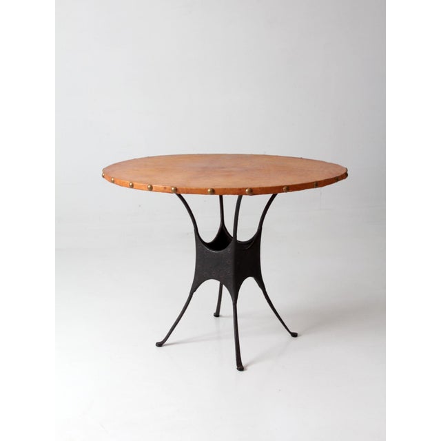 A vintage mid-century round dining table with a richly aged leather-covered top, sculptural iron base, and brass nailhead...