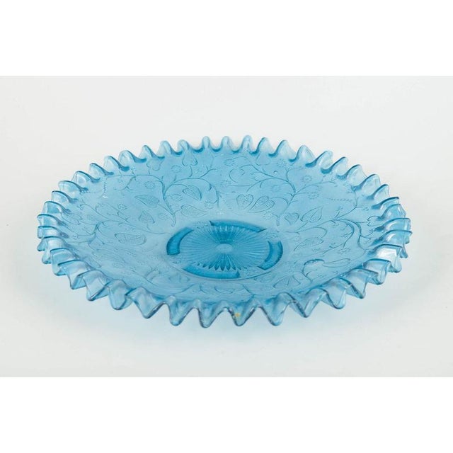 Decorative pressed-glass dish in light blue with a ruffled edge and an embossed floral and vine pattern. A characteristic...