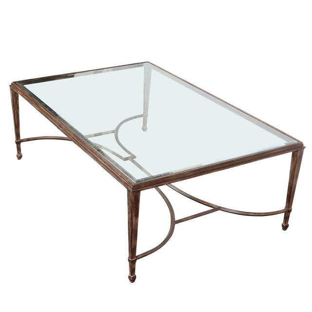 A large coffee table that features a gilt metal frame and a beveled glass top. The base is designed with a curved X-form...