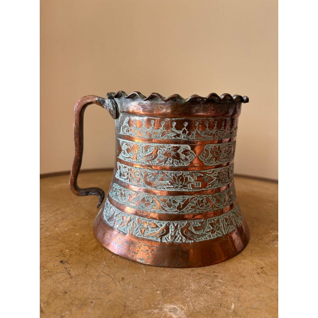 This Persian tankard is crafted from copper and tin. It features a fluted top and intricate, traditional floral and bird...