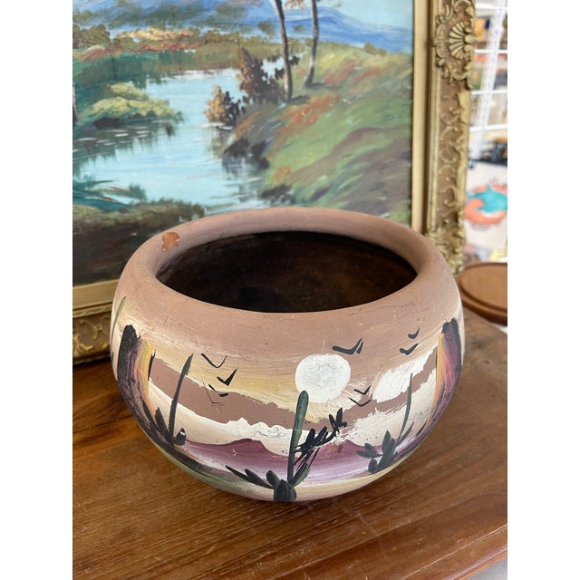 This vintage hand-painted terracotta pot features a vibrant desert landscape rendered in rich, earthy tones. The scene...