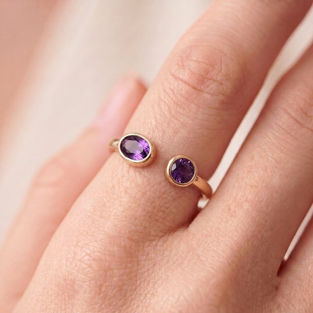 A bold, contemporary open-cuff ring featuring two vivid purple amethysts — an oval-cut stone paired with a round-cut stone...